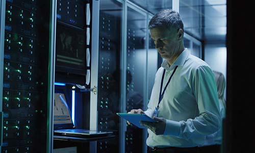 Man wearing a badge and button down shirt stands in front of a wall of technology equipment for software implementation.