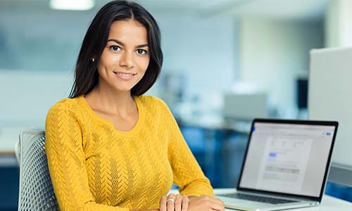 Woman in yellow shirt smiles next to a computer while handling the management of each phase of the project lifecycle.