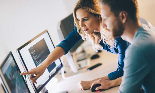 Woman points at computer while man sits at the desk, working together for quality assurance and function testing.